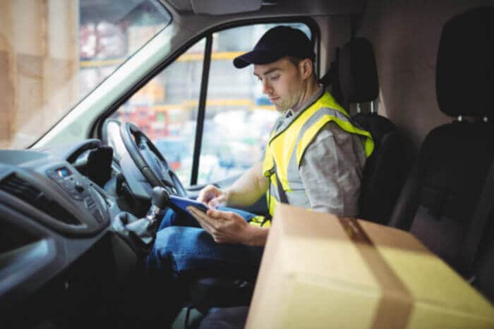 Delivery-Driver-Using-Tablet-In-Van-With-Parcels-On-Seat